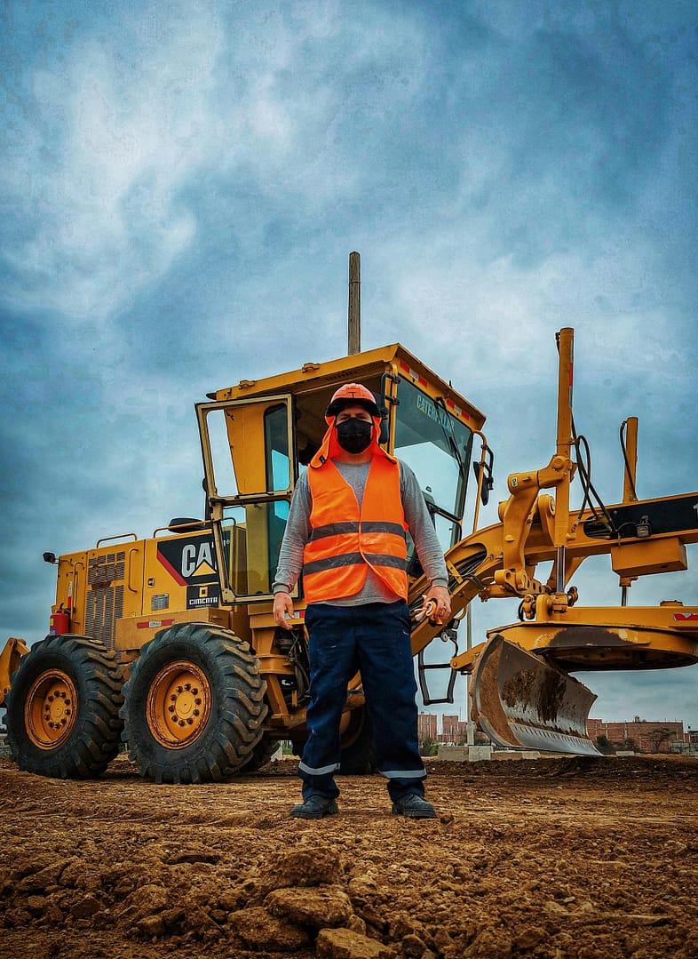A construction worker in safety gear stands in front of a bulldozer under a cloudy sky on a worksite.